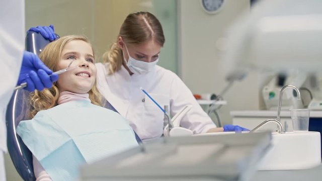 Cute little girl smiling while having dental procedure with dentist and assistant at the dental clinic