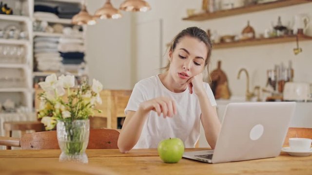 Bored young brunette woman holding an apple while waiting a letter on laptop computer sitting in the dining room
