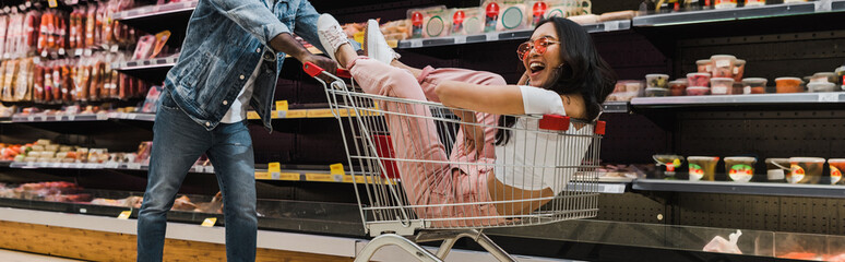 panoramic shot of happy asian girl in sunglasses sitting in shopping cart near african american man