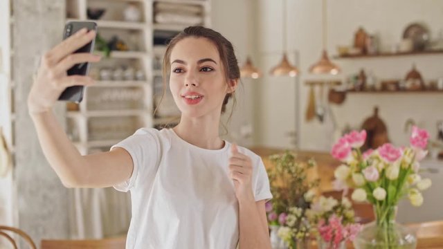 Cute young brunette lady in white t-shirt making a selfie photo on smartphone standing at the dining room