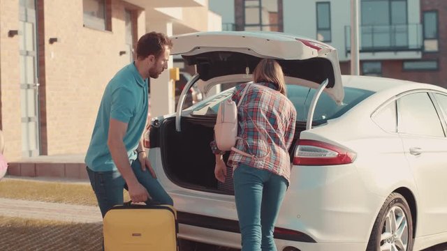 Beautiful traditional family packing for a summer vacation. Happy parents loading suitcases into a car trunk with help of their cute kids in the street.