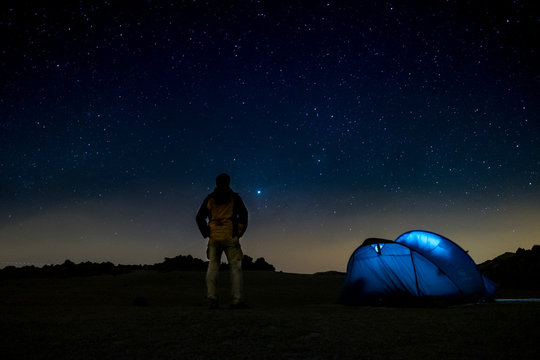 Night shot with dark blue sky and stars in background - concept of travel and freedom with tent camping in outdoor wild nature - standing man looking at the landscape around - adventure