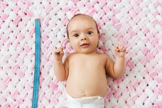 babyhood, size and high concept - sweet little baby girl in diaper lying with measuring tape on knitted pink blanket of plush yarn