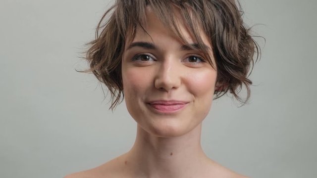 Shy young woman with natural skin smiling while looking at the camera over grey background isolated