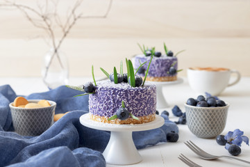 Two round no bake blueberry mini cheesecakes enveloped in coconut and embellished with real blueberries and rosemary leaves. White wooden table with coffe and milk in background.