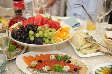 Naklejka na ściany i meble White and black grapes and sliced ​​watermelon in a plate, sliced ​​red and white fish with cheese and black olives in a plate. Wine glasses and wine. Festive table setting. Mediterranean. Catering.