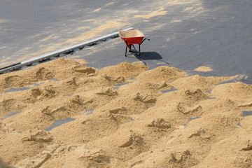 Fototapeta na wymiar Red wheelbarrow with one wheel on the pavement and many small slides of sand on the ground. Improve road paths in the city, work outside on a hot sunny day.