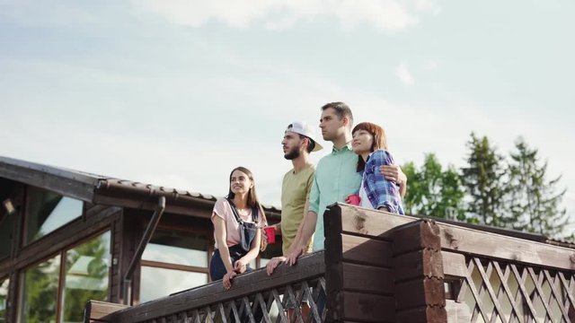 Camera moving away from four young friends, two couples, standing at railing of wooden countryside house terrace, looking at view and discussing nature