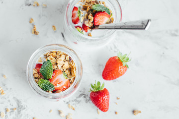 Two jars with tasty parfaits made of granola, strawberries and yogurt on white marble table. Top view, copy space