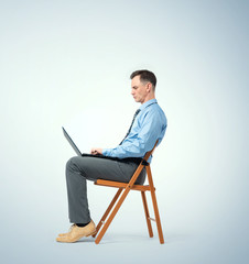 Man in a blue shirt with a tie sits on a chair working with a laptop.