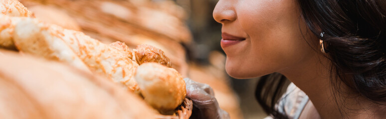 panoramic shot of cheerful woman smiling while smelling bread in supermarket