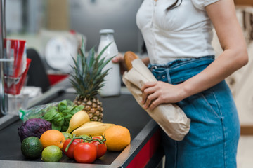 cropped view of girl holding baguette and glass bottle with milk near supermarket counter