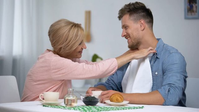 Careful mother putting napkin on son neck during having tea, overprotection