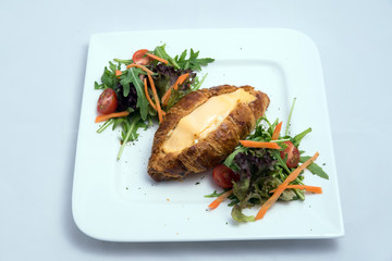 A low contrast image of a breakfast platter with cheese-filled croissant with vegetables- carrot, tomato, lettuce on a minimal white background with a 30 degree angle from front zoomed in perspective