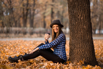 Fall concept. Happy and cheerful woman in hat, drinking coffee while sitting on park leaves under fall foliage. Beautiful young modern woman smiling