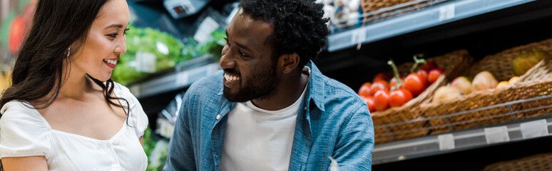 panoramic shot of happy african american man looking at cheerful asian girl in store