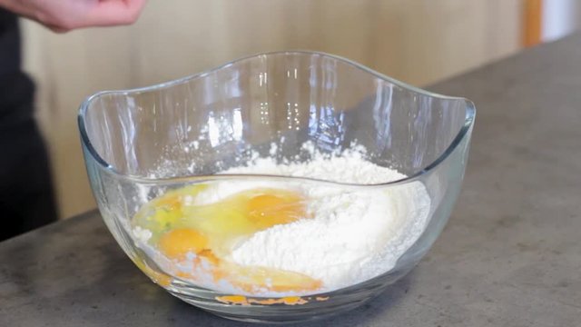 The guy breaks the egg in a round, glass, transparent plate, which is flour and other yolks of eggs. Cooking. The guy cooks food in the kitchen.