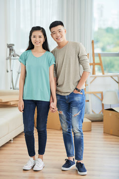 Portrait of young Asian couple standing in casual clothing holding hands and smiling at camera at home