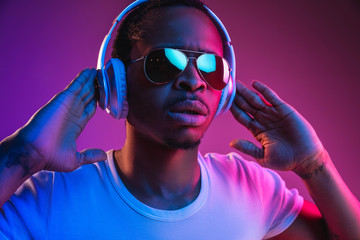 Young african-american man's listening to music in headphones in neon light on gradient background....