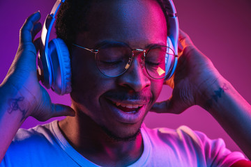 Young african-american man's listening to music in headphones in neon light on gradient background....