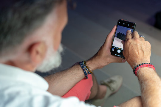 Old man with white beard touching the screen of his smartphone at summer night