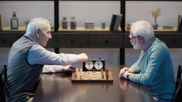 side view of two pensive senior men sitting at table, playing chess and pressing button on chess clock in living room