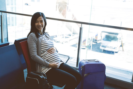 asian pregnant woman sitting beside a suitcase on the airport waiting room before flight departure