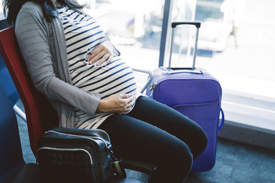 asian pregnant woman sitting beside a suitcase on the airport waiting room before flight departure