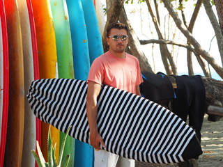 Portrait of a male surfer in a fashionable t-shirt holding a surfboard at the surfsport. Background bright surfboards.