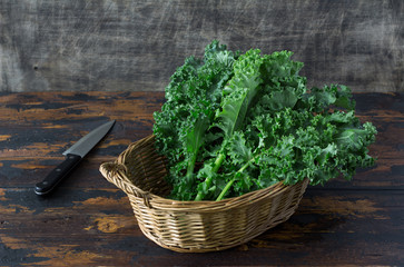 Fresh organic curly kale leaves in a basket with cook knife behind, on a brown wooden table and on...