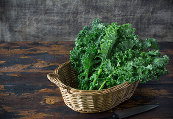 Fresh organic curly kale leaves in a basket with cook knife in a front, on a brown wooden table and on a dark wooden background, gmo free,  rustic style, copy space, horizontal board, free space left - obrazy, fototapety, plakaty