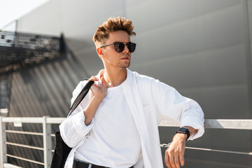 Cool young hipster man in a white t-shirt in a stylish shirt in trendy sunglasses is standing near...