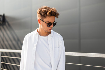 Stylish young man model in vintage white shirt in trendy sunglasses with a hairstyle stands near a gray building on a sunny day. European hipster guy on the street. New summer collection menswear.