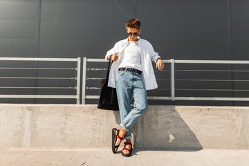 Modern young man hipster in fashionable summer white clothes in sunglasses in stylish red leather...