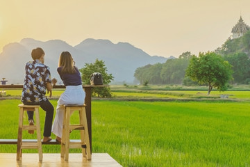 Back view of couple sit for resting and waiting for time to take photos of the sunset on the farmer's balcony in the rice fields in front of Wat Tham Sua in Kanchanaburi, Thailand.