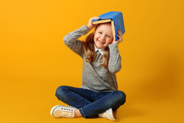 Cheerful attractive little student girl is sitting on the floor with a book on her head. The...