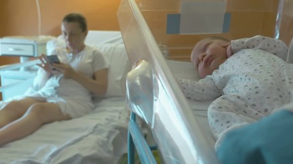 Newborn baby sleeps in a medical baby cradle, a young mother after giving birth lies on the hospital cot and looks into the phone on the background. Cute newborn baby smiling in his sleep