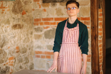 Young male artist in aprong and glasses stand in front of brick wall