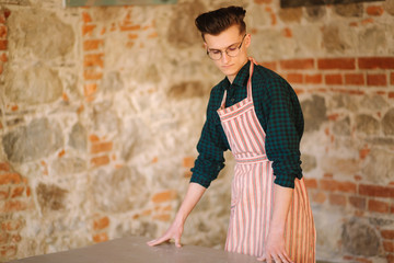 Young male artist in aprong and glasses stand in front of brick wall