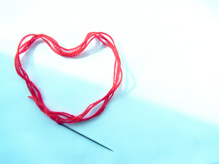 A red woolen thick thread with a sewing needle lies in the shape of a heart, lit on the white and blue background.