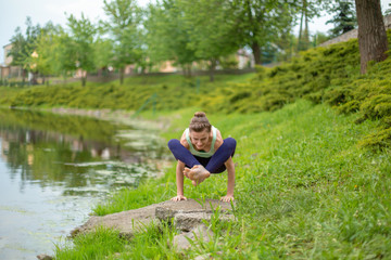 Naklejka na ściany i meble Thin brunette girl plays sports and performs beautiful and sophisticated yoga poses in a summer park. Green lush forest and the river on the background. Woman doing exercises on a yoga mat
