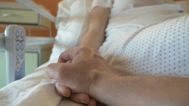 Man holds the hand of the woman who lies in the modern hospital cot. Support for a sick person in the ward, patient after surgery feels pain. Emotional moment in close-up