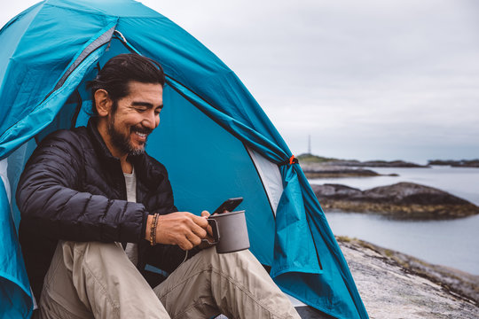 Man with a mug and a cellphone sitting at a camping tent by the sea