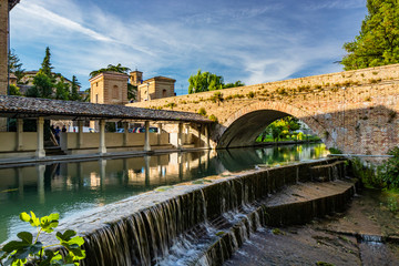 Fototapeta premium The ancient wash-house and the masonry bridge over the river, in the medieval village of Bevagna. Perugia, Umbria, Italy. Blue sky at sunset. Trees and vegetation. The reflection on the water surface.
