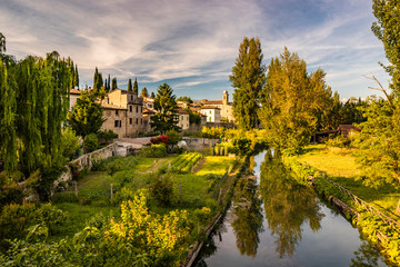 Fototapeta na wymiar The Chiasco river that crosses the ancient medieval village of Bevagna. Perugia, Umbria, Italy. Trees, vegetation, cultivated gardens. The blue sky at sunset, in the summer.