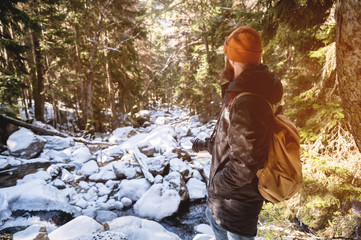 A bearded man in a hat and a backpack stands with his back in winter in a coniferous forest on the river. The concept of winter travel and walks in the woods