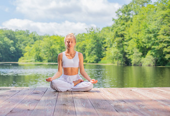 Fototapeta na wymiar Attractive woman is practicing yoga and meditation, sitting in lotus pose near lake in morning.