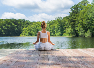 Attractive woman is practicing yoga and meditation, sitting in lotus pose near lake in morning.