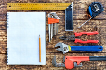 Various hand tools and blank notepad with pencil on old wooden background.