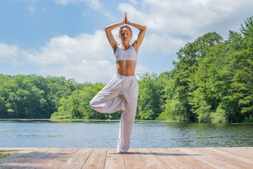Attractive woman is practicing yoga, doing Vrksasana exercise, standing in tree pose near lake.